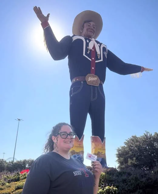 A woman with glasses poses, holding an Irving Library card in front of a statue of Big Tex at the State Fair.