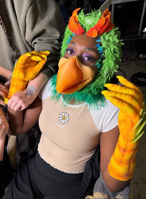 The rapper Noname in a prosthetic beak and rubber bird hands with a green feathered hood smiles at camera. A makeup artist leans into frame.