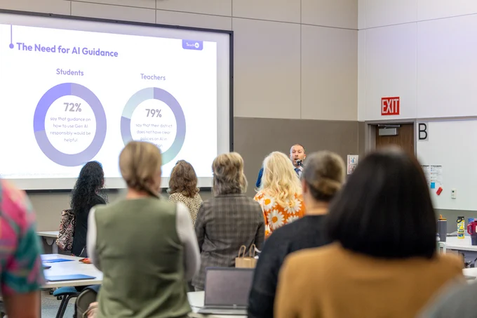 A conference room with attendees facing a presenter near a screen displaying pie charts and the title "The Need for AI Guidance."