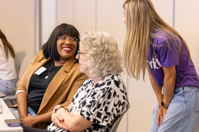 Three individuals in a meeting room, engaging in a discussion. One person is wearing a tan blazer and black glasses, another in a black and white patterned top, and the third in a purple t-shirt. They appear to be sharing a joyful conversation.