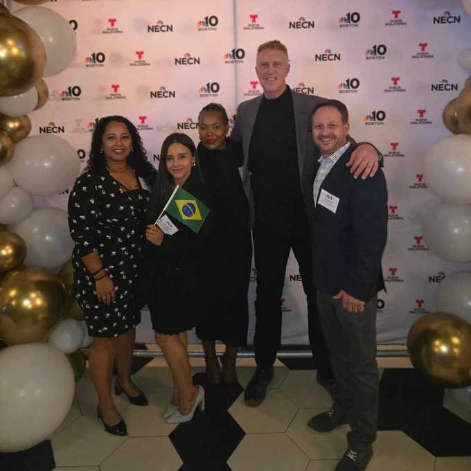 Five people in semiformal outfits between balloons: two Latina women, a Black woman, and two White men. They're smiling in a posed picture in front of a NBC10, Telemundo, and NECN backdrop. One woman is holding a small Brazilian flag.