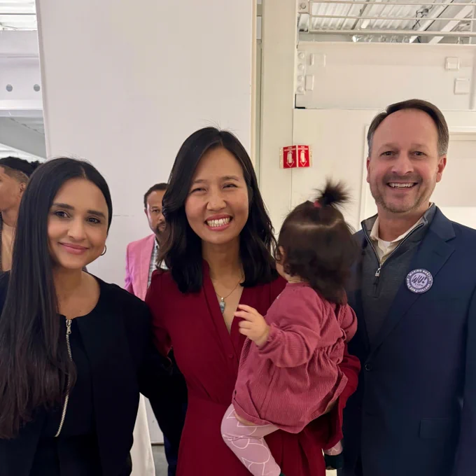 A Brazilian woman with long, dark hair and a white man with short brown hair stand on either side of Mayor Michelle Wu and smile. Mayor Wu is smiling and holding her child, who is facing away from the camera.