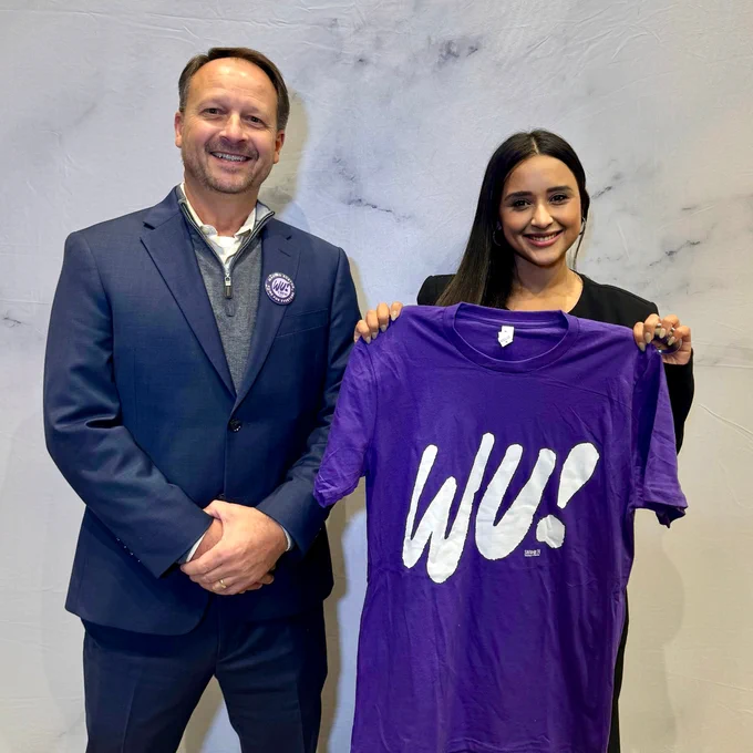 Two people, a white man with short hair wearing a blazer and vest and a Brazilian woman holding a purple Mayor Michelle Wu t-shirt smile in a posed photo.