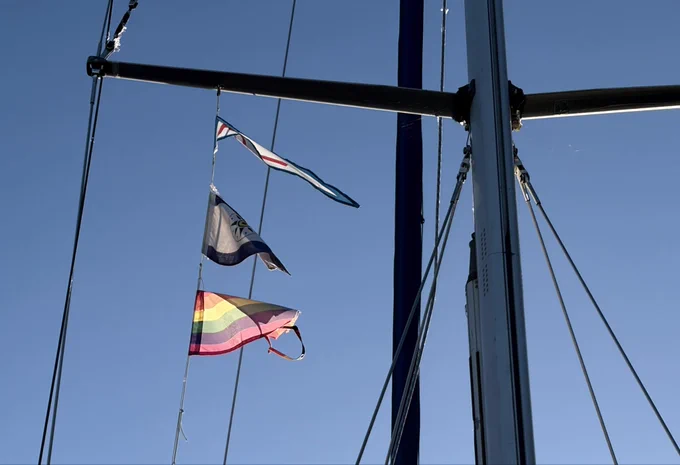 Drei Flaggen im Rigg eines Segelbootes vor blauem Himmel. Im oberen Bildteil sieht man ein Salingspaar, das sind quer zum Mast angebrachte Stangen. Rechts im Bild der Mast und das aufgerollte Vorsegel. Von oben nach unten durch das Bild laufen die Drähte, die den Mast halten. Unter der Backbordsaling wehen ein Vereinsstander, die Flagge des schwedischen Kreuzerklubs sowie eine Pride Flag. Letztere ist ausgerissen. Der Himmel ist SEHR blau.