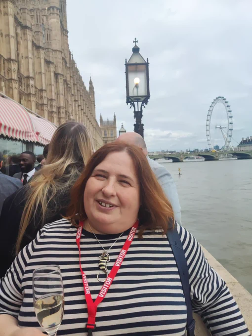 Susan holding a glass of champagne, standing on the river terrace at the Houses of Parliament with the London eye in the background.