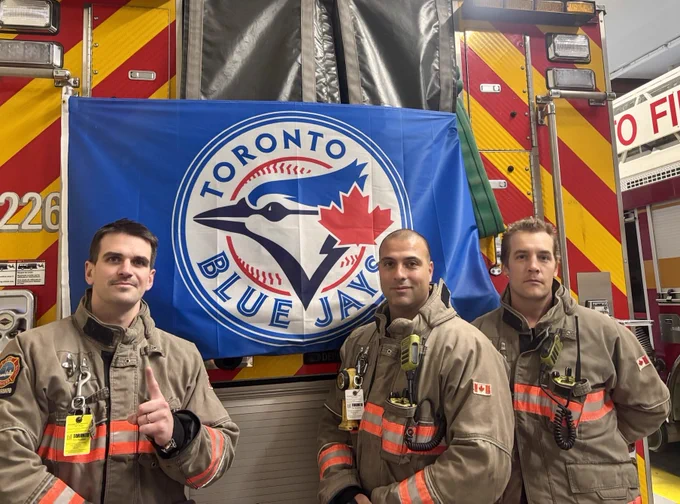 Three Toronto Fire Services Operations Firefighters in bunker gear standing in front of the back of a fire truck with the Toronto Blue Jays flag hanging off the back of the fire truck.