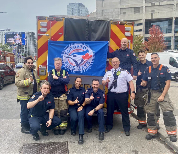 Toronto Fire Services crew members in blue fatigues and bunker suits standing in front of the back of a Toronto fire truck. They people are holding up their index finger to symbolize #1 and one crew member is holding a Toronto Blue Jays flag.