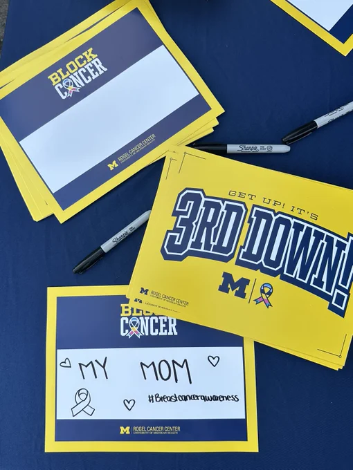 Signs and markers on a blue table for a University of Michigan cancer awareness event, featuring slogans like "Block Cancer" and "3rd Down!" along with a personal message reading "My Mom" to promote cancer awareness.
