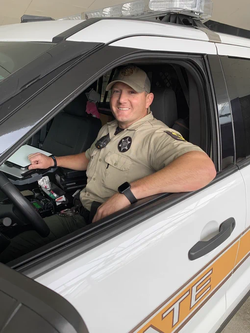 A law enforcement officer in a brown uniform, smiling from the driver's seat of a patrol vehicle.