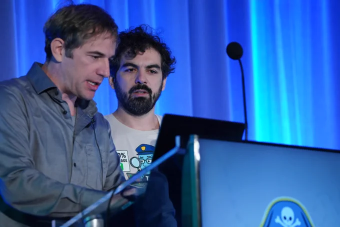 A man with curly dark hair and a beard is speaking on a stage against a bright blue curtain backdrop, standing behind a podium with a laptop. He is wearing a light-colored t-shirt with a graphic design of a figure on it. He is gesturing with his left hand and looking directly forward with a serious expression.