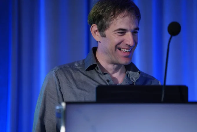 Two men are standing behind a podium with an open laptop, against a bright blue curtain backdrop. The man in the foreground, wearing a gray button-down shirt, is looking down and to the left with his mouth slightly open as if speaking. The man behind him, with curly hair and a beard, is wearing a light-colored t-shirt with a graphic on it and is looking directly at the camera.