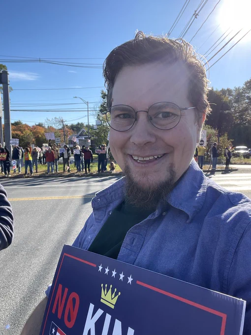 Bethany Andres-Beck smiles holding a “N0 KINGS” sign in Rowley, MA. Across the street is a crowd of protesters with signs and American flags.