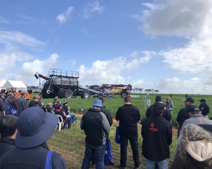 A group of people attending an outdoor agricultural demonstration, focusing on a large farming sprayer being displayed in a field.
