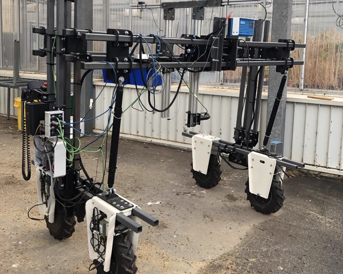 A complex agricultural robot equipped with various sensors and electronic devices, standing in a greenhouse environment.