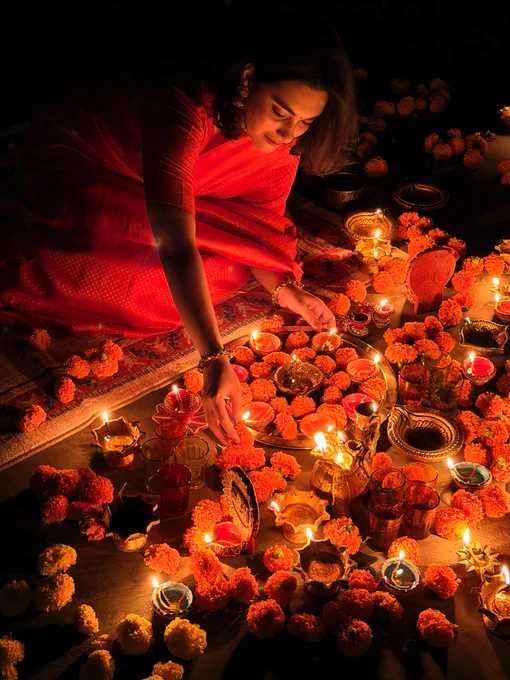 A woman dressed in a red saree kneels on a carpet and arranges a display of marigold flowers and lit diyas in front of her.