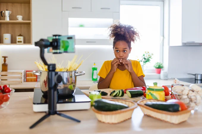 Young in a kitchen with fresh vegetables and pasta on the counter eats while a cell phone films her.
