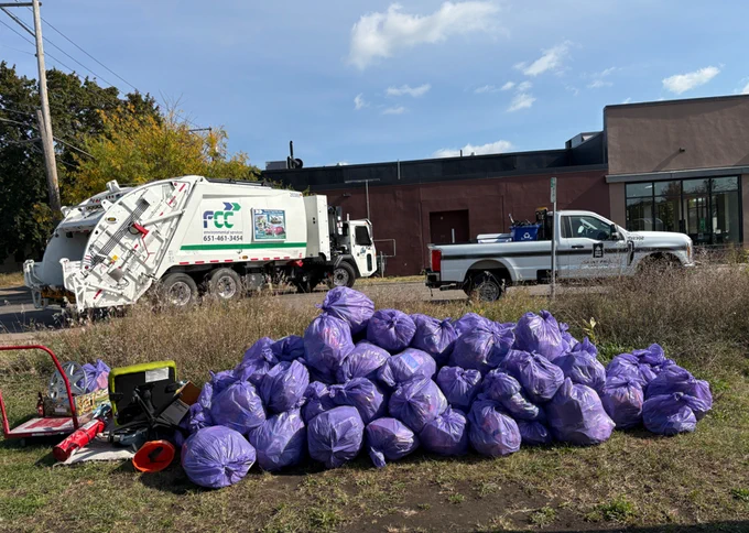 Photo credit: HMC from their litter clean up event on 10/18/25. Photo shows A large pile of purple trash bags stacked on a grassy area in front of a white garbage truck and a white utility pickup truck. The truck has branding and a phone number on its side. A brown building and power lines are in the background under a clear, blue sky. Nearby, there's various equipment, including bins and a red wagon.