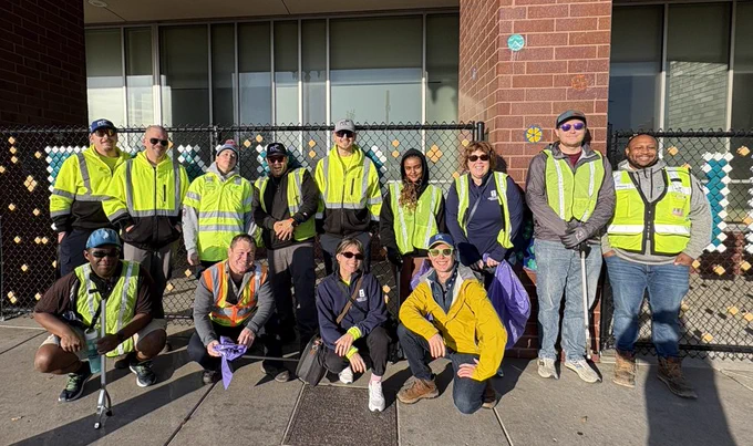 Photo of city staff and volunteers doing a cleanup event on 10/18/25. Photo shows: A diverse group of people wearing yellow and green reflective safety vests pose in front of a brick building and chain-link fence. Some are kneeling while others stand, smiling at the camera. The morning sunlight casts long shadows, creating a bright and lively atmosphere.