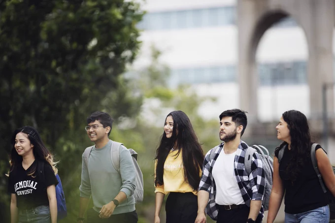 A group of Memorial University students walk outdoors on the St. John's campus. They are smiling and chatting while wearing backpacks. Two students are wearing 'Memorial University' shirts.