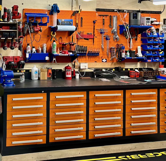 A neatly organized workshop featuring bright orange Wall Control Metal Pegboards mounted above a black workbench with orange drawers, holding various tools, bins, and supplies in a clean and efficient layout.