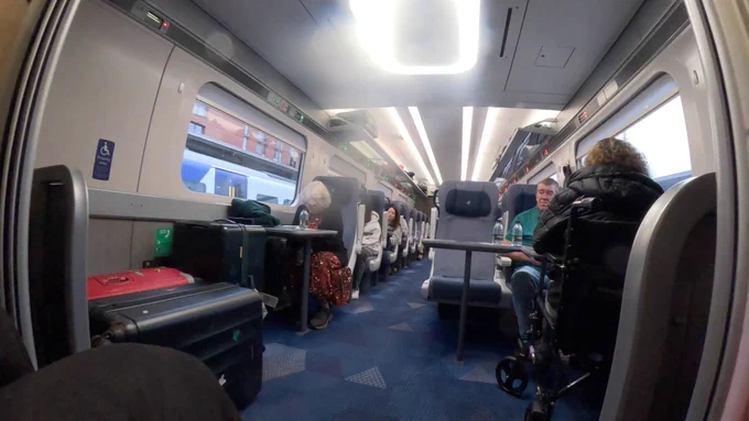Entrance to a train coach, from my camera attached to my wheelchair. 
One wheelchair space with wheelchair user in with her back to me, the other, clearly marked wheelchair space with reservation sign, has giant suitcases in it. 