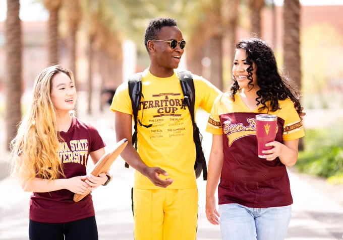 Three college students walk together outdoors on a sunny day, wearing Arizona State University apparel and carrying a tablet and a tumbler with the ASU Pitchfork logo.