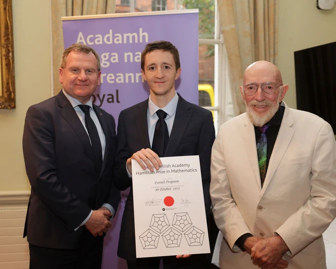 Three men in suits stand together. The man in the center, holding a certificate titled "Royal Irish Academy Hamilton Prize in Mathematics" dated 10 October 2023, smiles slightly. The backdrop features a purple banner with Irish text.