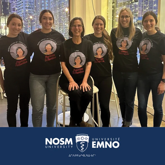 Six women are in a line, wearing matching black t-shirts. On the image on the t-shirts is a drawing of Dr. Frances Kilbertus, who herself appears in the photo, third from left in the line.
