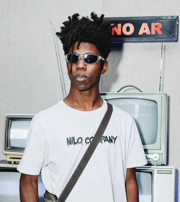 Abelha, um homem preto com cabelo preto em locs, vestindo uma camiseta branca, uma bolsa verde desbotado com a alça atravessando seu ombro e óculos escuros com armação cinza. Ele tem expressão neutra e se posiciona na frente de televisões de tubo antigas, com uma placa preta com texto vermelho acima que se lê: "No ar".