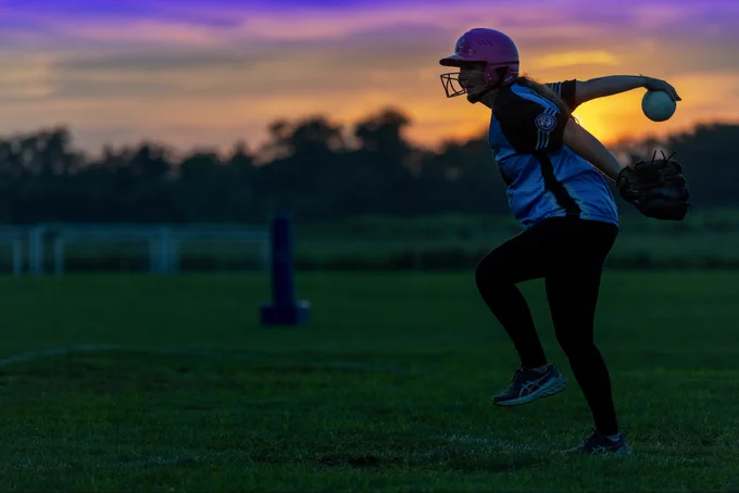 Molly Fleming, pitching for the Southwest Bombshells, winds up to pitch the ball. She wears a pink helmet and a blue jersey with black sleeves, her right arm extended back holding the ball. The sky glows with a vivid sunset of orange and purple hues behind a line of trees, creating a dramatic backdrop over the green field.