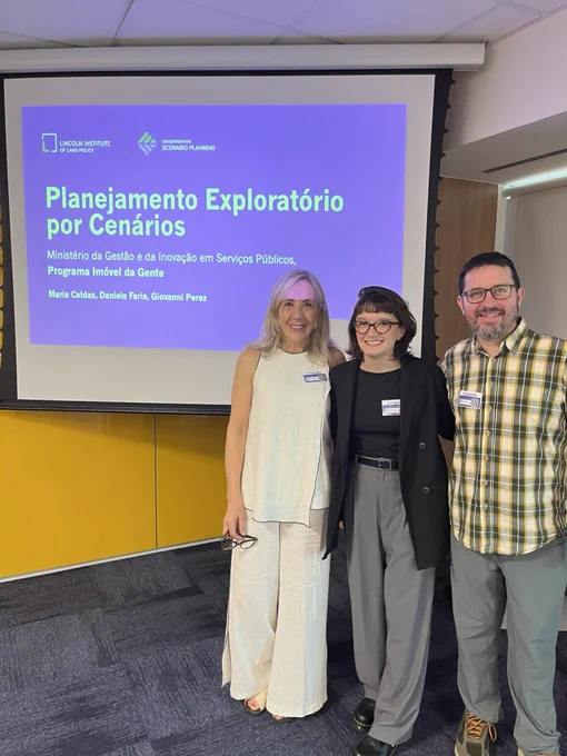 Three presenters stand smiling in front of a projected slide in a conference room. All wear name badges. The slide shows the Lincoln Institute of Land Policy and Consortium for Scenario Planning logos and reads “Planejamento Exploratório por Cenários,” with subtext “Ministério da Gestão e da Inovação em Serviços Públicos, Programa Imóvel da Gente.” The slide lists the presenters’ names: Maria Caldas, Daniela Faria, and Giovanni Perez.