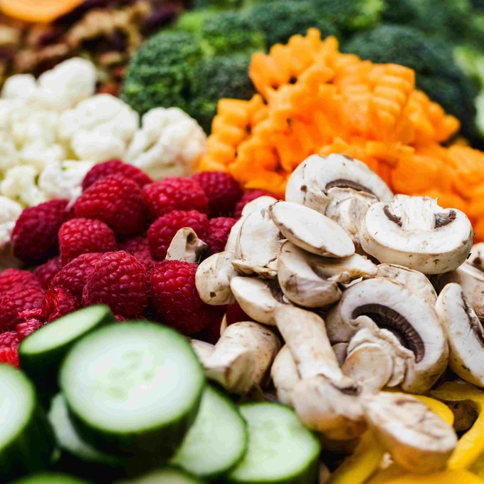 Assorted fresh vegetables and fruits, including broccoli, carrots, cauliflower, raspberries, mushrooms, cucumbers, and bell peppers, arranged in a vibrant display.