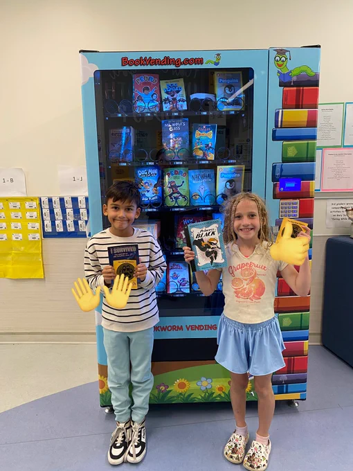 Two students stand with books in front of a book vending machine.