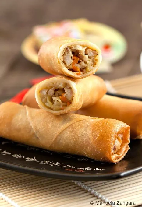 Golden fried chicken spring rolls stacked on a black plate with dipping sauce in the background.