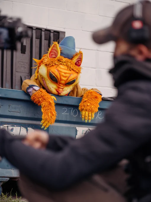 A cat like anthropomorphic creature emerges from a dumpster. In the foreground, a camera person had to focus crouches to record the moment.
