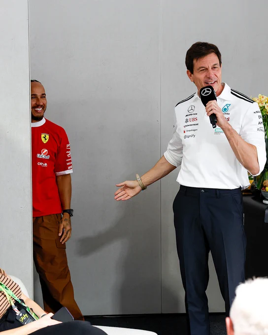 Toto Wolff holding a microphone and pointing towards Lewis Hamilton who is peeking out from behind a wall in hospitality.