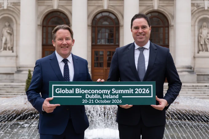 Two individuals holding a sign that reads "Global Bioeconomy Summit 2026 Dublin, Ireland 20-21 October 2026" in front of a building with a fountain.