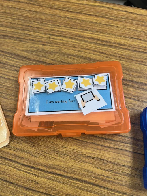 orange plastic box with laminated sheet on outside "I am working for" with a space for a preferred item.  four spaces for tokens to stick on velcro