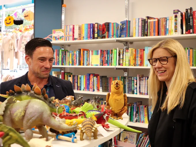 Two adults smiling and looking at a collection of colourful dinosaur toys and a plush toy on a table in a bookstore.