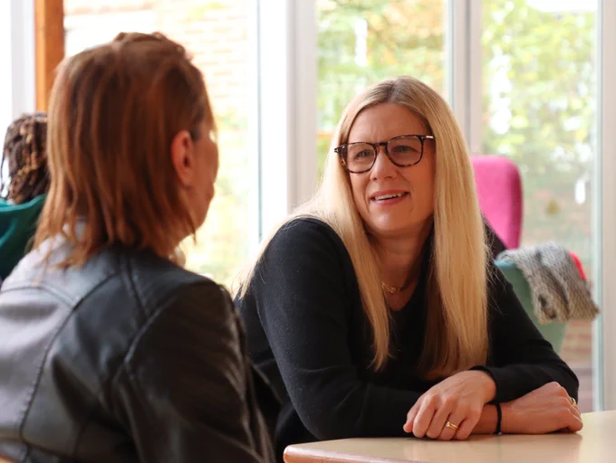 Two people having a conversation at a table with a bright window in the background. One individual is facing the camera and is smiling.