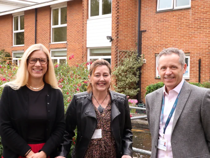 Three individuals (Ellie Reeves MP, Steve Smith, and a hospice service user) standing in front of a building, smiling, with a garden and a small fountain in the foreground.