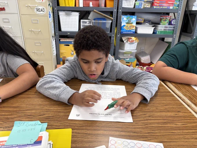 A close-up of an elementary school-aged boy with curly hair, wearing a grey hooded sweatshirt, concentrating on a worksheet titled "The Fox and the Red Hen." He is holding a green marker over the paper. The edge of a binder labeled "3rd Grade" is visible in the foreground.