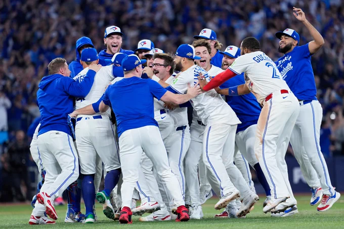 Members of the Toronto Blue Jays celebrate at the mound following the Blue Jays clinching the American League pennant. They all wear white Blue Jays uniforms with blue lettering and white and blue caps.