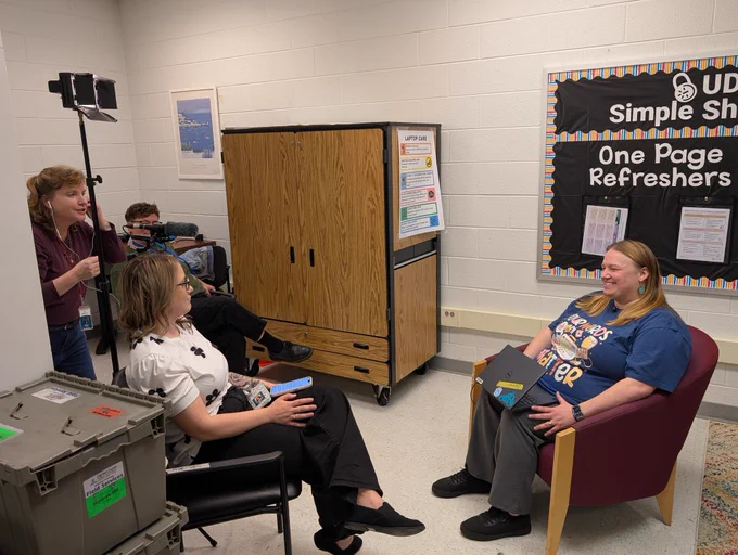 Three adults with video equipment are on the left side of a room. An adult female is sitting on the right and is being interviewed.