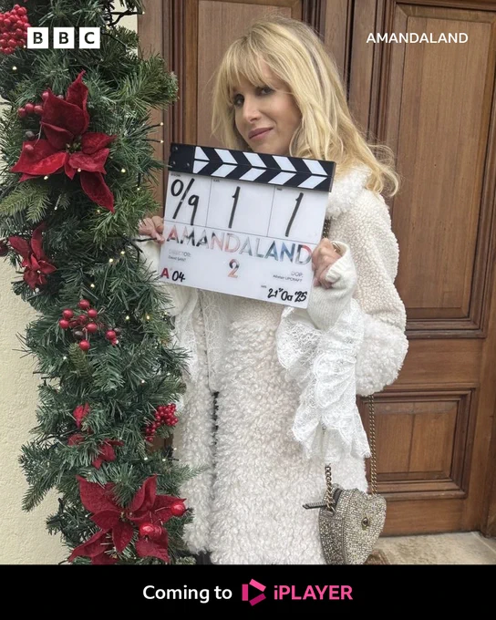 A woman in a white jacket stands outside a wooden door, decorated with festive greenery and red ribbon, holding a clapperboard for the start of filming on Amandaland. Text in white reads Coming to iPlayer.