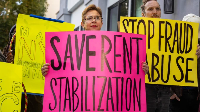 A tenant advocate holds a pink poster reading "Save rent stabilization"