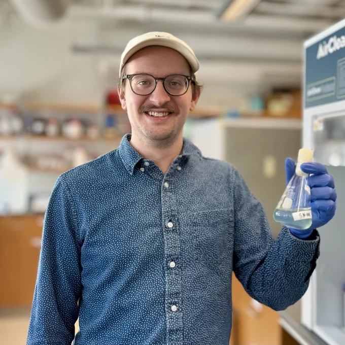 Headshot of Dr. Henry Holm. Henry holds an Erlenmeyer flask containing a phytoplankton culture while working in a lab at Lamont-Doherty Earth Observatory. Photo by Henry Holm. 