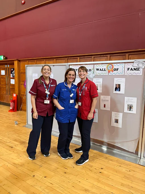 Three healthcare workers in scrubs, each wearing a badge, standing together in a room with a "Wall of Fame" banner in the background.