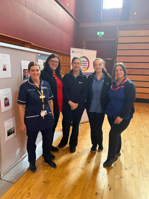 Five healthcare professionals smiling in a well-lit room, standing next to an informational display. They are wearing different uniforms, signifying varied roles.