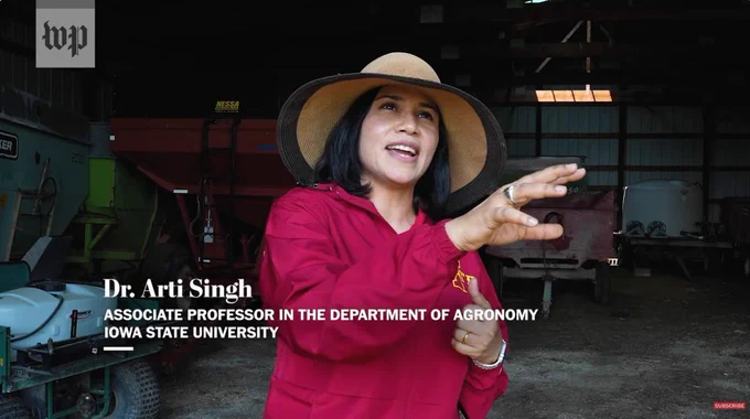 Dr. Arti Singh wearing a broad brimmed hat talking to the camera while standing next to farm implements.